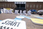Kiss Kiss Way 2025: Villaggio con palco, stand brand e pubblico festante durante una tappa estiva del tour