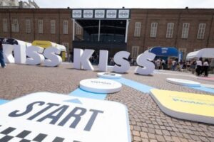 Kiss Kiss Way 2025: Villaggio con palco, stand brand e pubblico festante durante una tappa estiva del tour